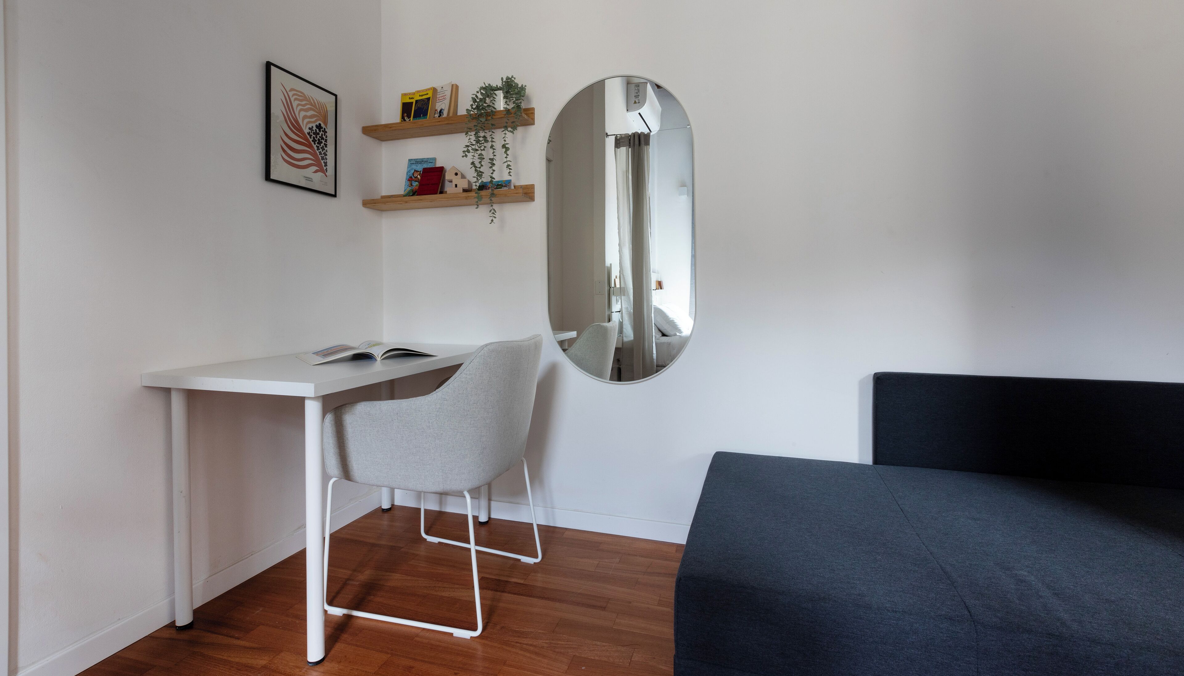 A compact room with a white desk, a grey chair, a large mirror, and a dark sofa.