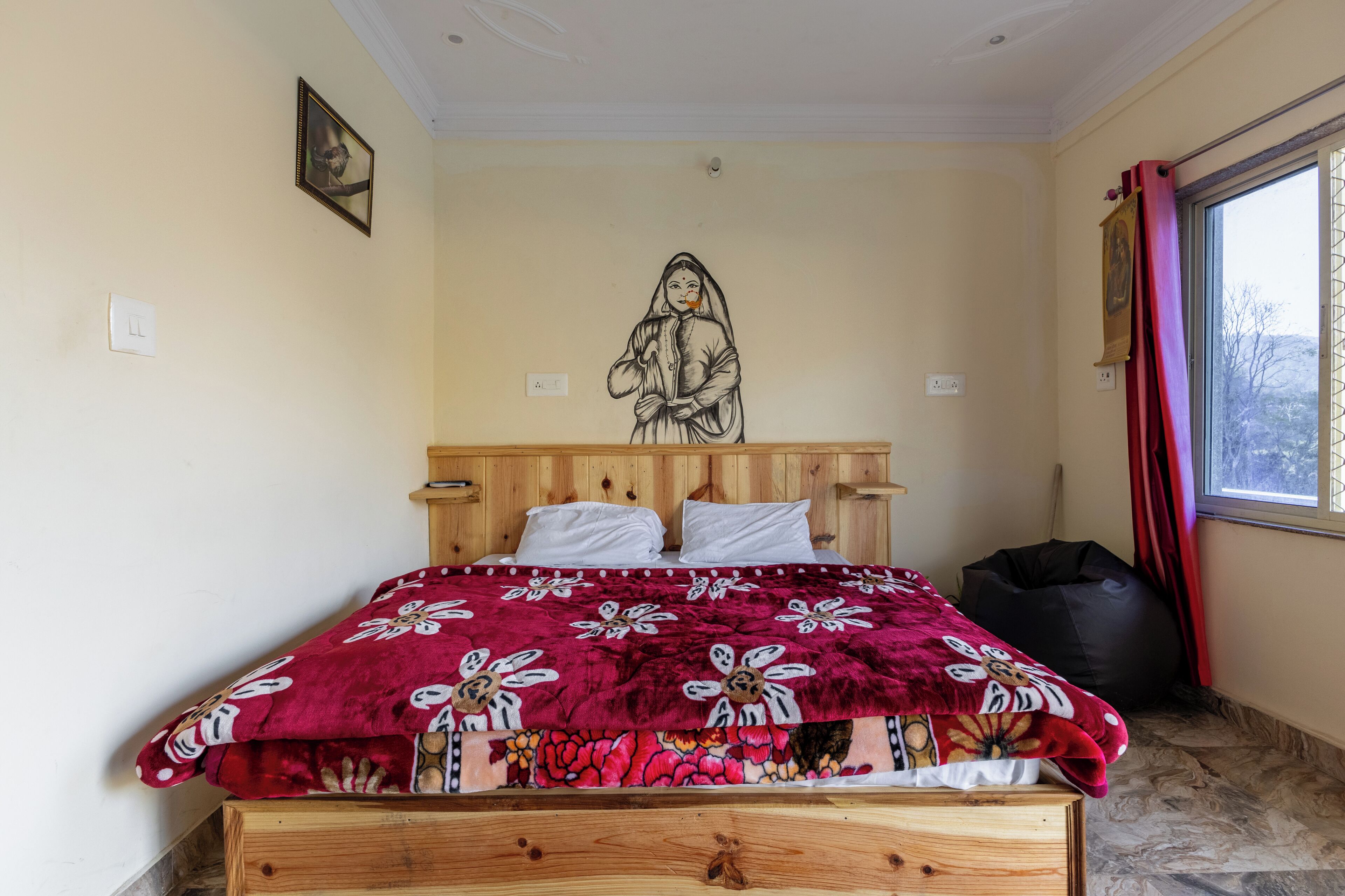 A bedroom with a wooden bed, a red floral bedspread, a black bean bag, a wall-mounted picture, and a window with red curtains.