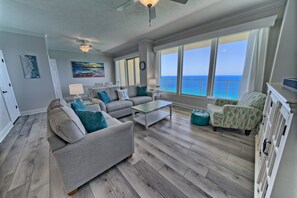 A spacious living room with a gray sofa, teal cushions, a coffee table, and a view of the ocean through large windows. - Gulf Crest 2305 | Beachfront Penthouse with Stunning Views (Panama City)