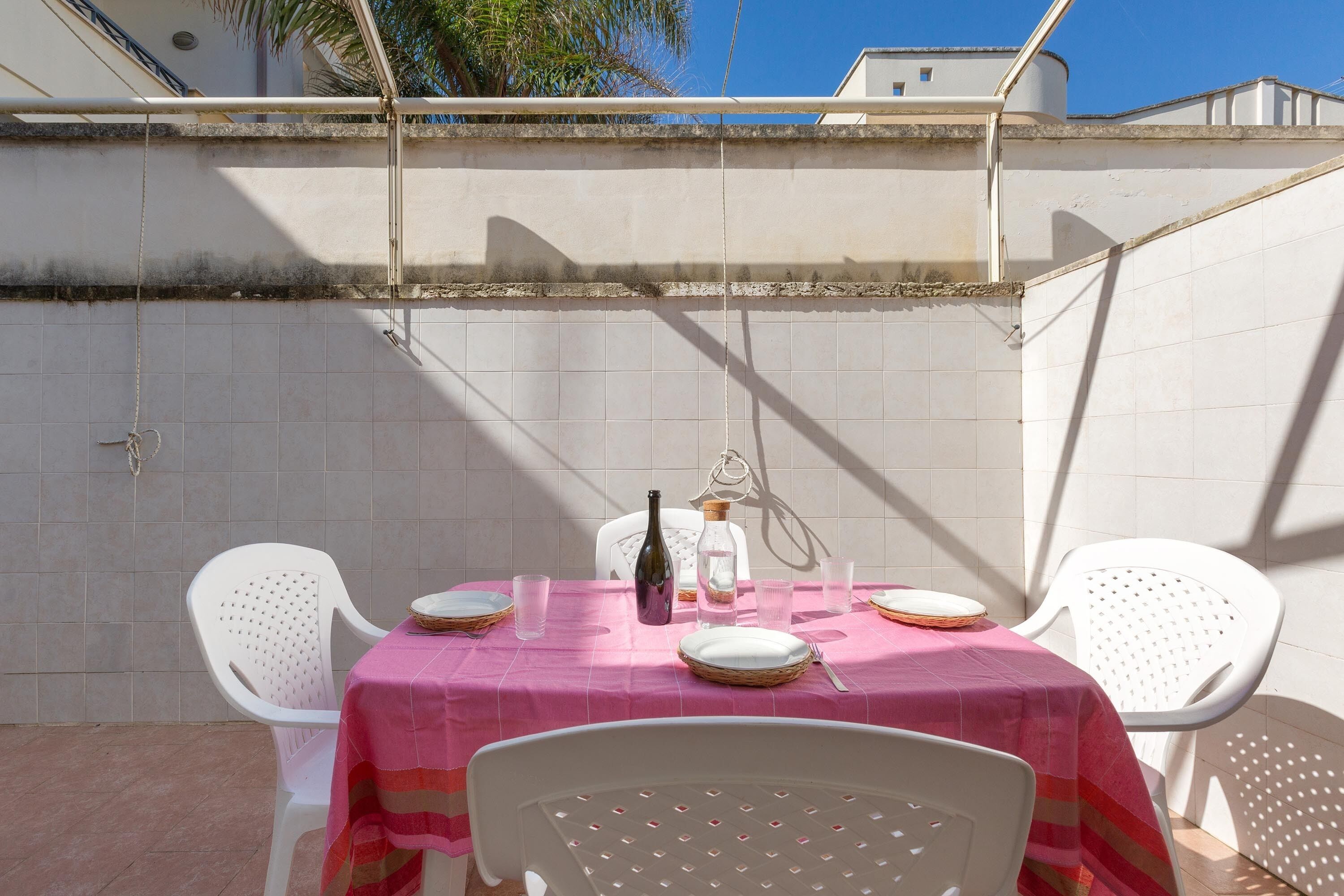 Eine Terrasse mit Tisch für zwei Personen, weißen Plastikstühlen und einer pinkfarbenen Tischdecke.