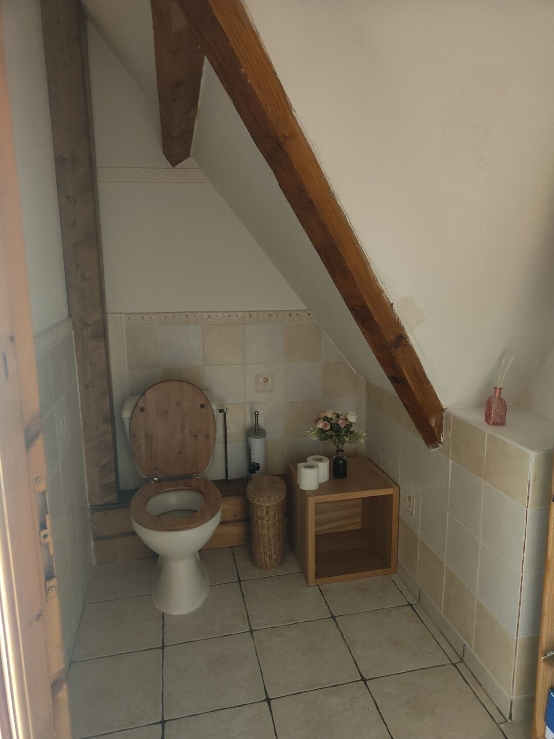 Une petite salle de bain rustique avec des toilettes en bois, une petite étagère contenant des articles de toilette et un sol carrelé.