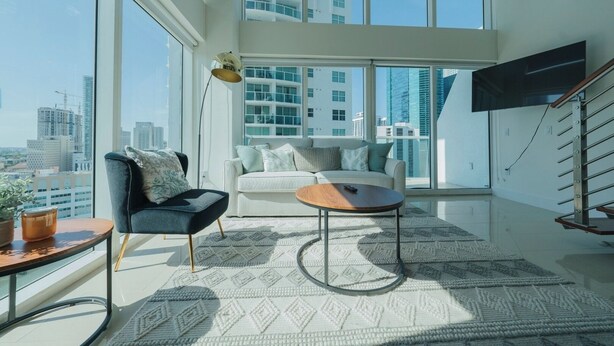 A modern living room with a sofa, armchair, coffee table, and a large window offering a city view.