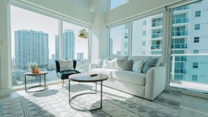 A spacious living room with a large window, a white sofa, a black armchair, and a round coffee table.