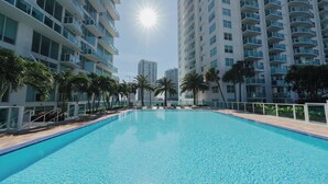 A swimming pool surrounded by high-rise buildings, palm trees, and a clear sky.
