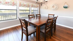 Une salle à manger avec une table et des chaises en bois, de grandes fenêtres ornées de stores à motifs floraux et un lustre.
