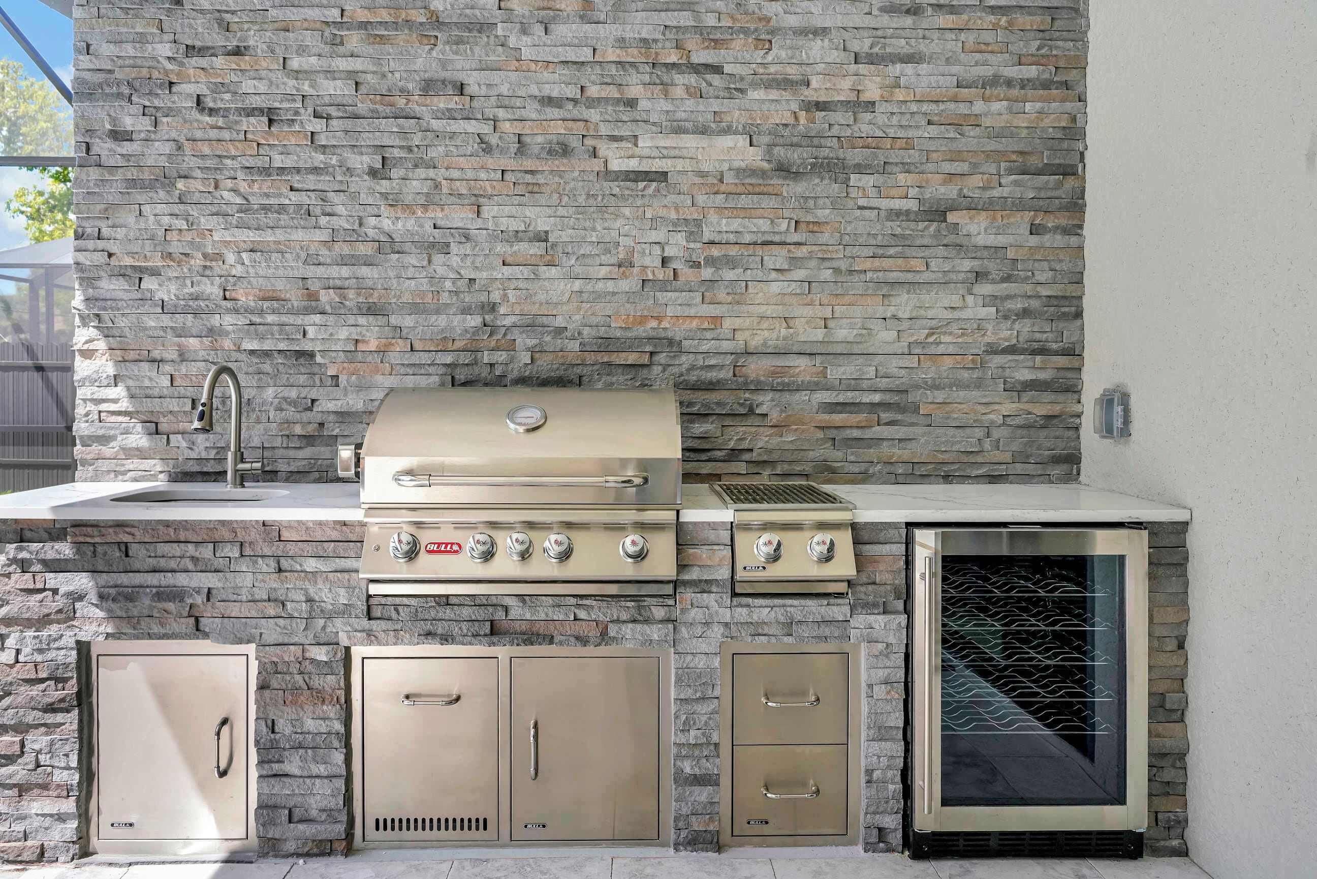 An outdoor kitchen with a stone wall backdrop, a stainless steel grill, a sink, and storage cabinets.