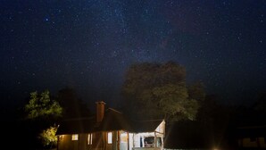Ein rustikales Blockhaus mit einem strohgedeckten Dach, beleuchtet von Innenlichtern, vor einem sternenübersäten Nachthimmel.