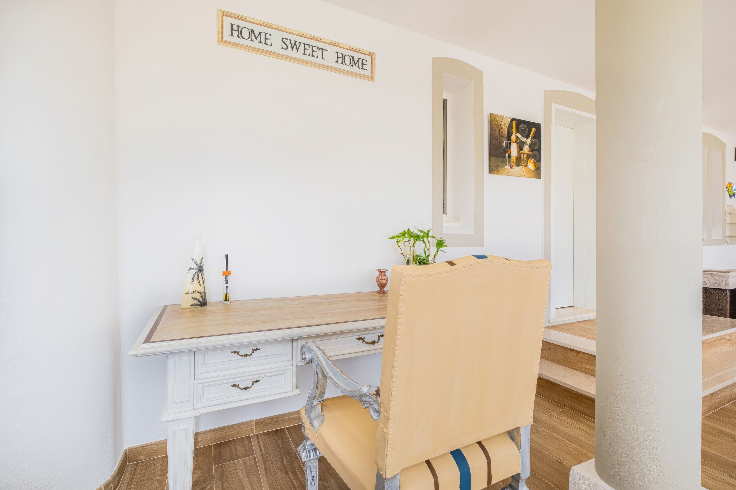 Et værelse med et hvidt skrivebord, en stol med en stribet pude, et skilt på væggen med teksten 'HOME SWEET HOME' og et indrammet billede.