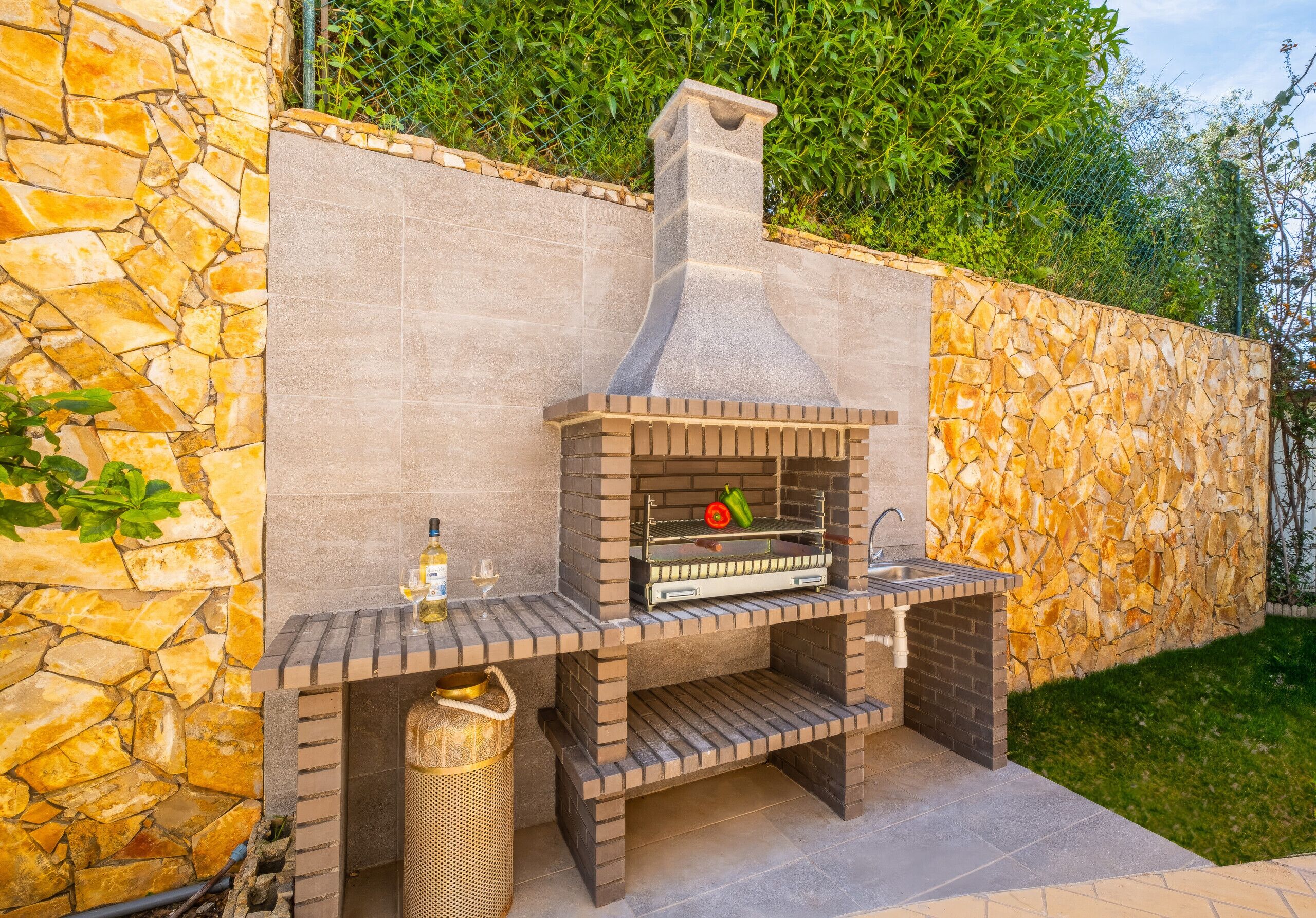 Zona de barbacoa al aire libre con horno de ladrillo, pared de piedra, pileta y una botella de vino.