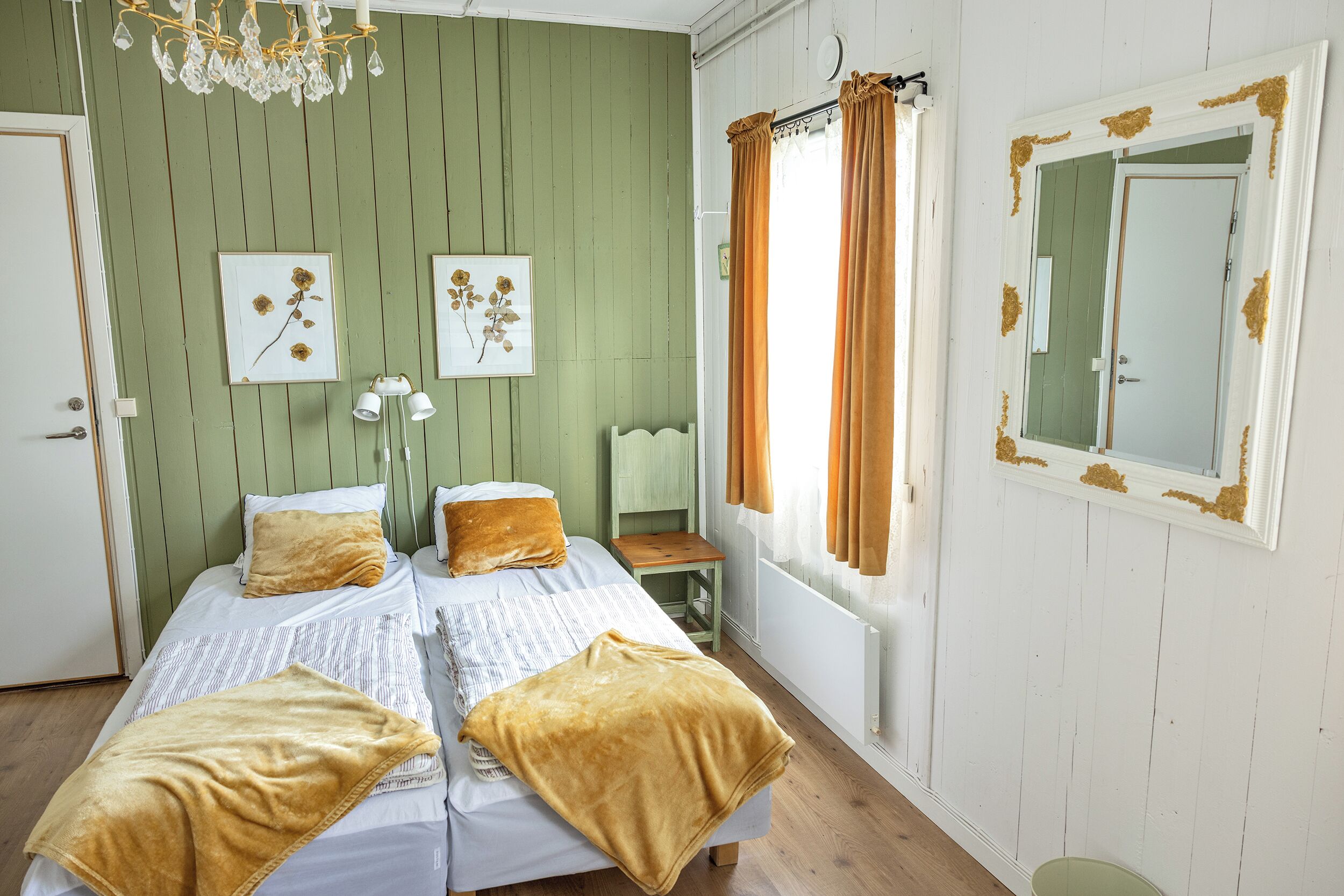 Une chambre avec deux lits, un lustre, des tableaux encadrés, une fenêtre avec des rideaux et un miroir orné de motifs décoratifs.