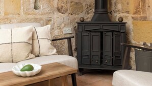 Una habitación acogedora con pared de piedra, un estufa de leña negra, un banco acolchado y una mesita con un cuenco.