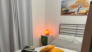 A neatly made bed with a white knitted cover, a green nightstand with a lamp, and a painting of a tree on the wall. - House in Silius R5130 (Silius)