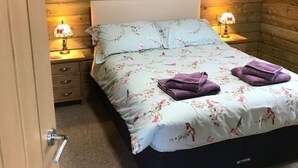 A bedroom with a wooden ceiling, a bed with floral bedding, two bedside lamps, and a nightstand with a lamp. - The Stables - Stokeinteignhead (Torquay)