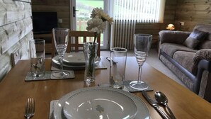 A dining table set with plates, glasses, and cutlery, a vase with flowers, and a view of the outdoors through a window. - The Stables - Stokeinteignhead (Torquay)