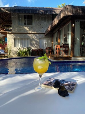 Une scène de bord de piscine : une boisson dans un verre, un livre et des lunettes de soleil sont posés sur une table.