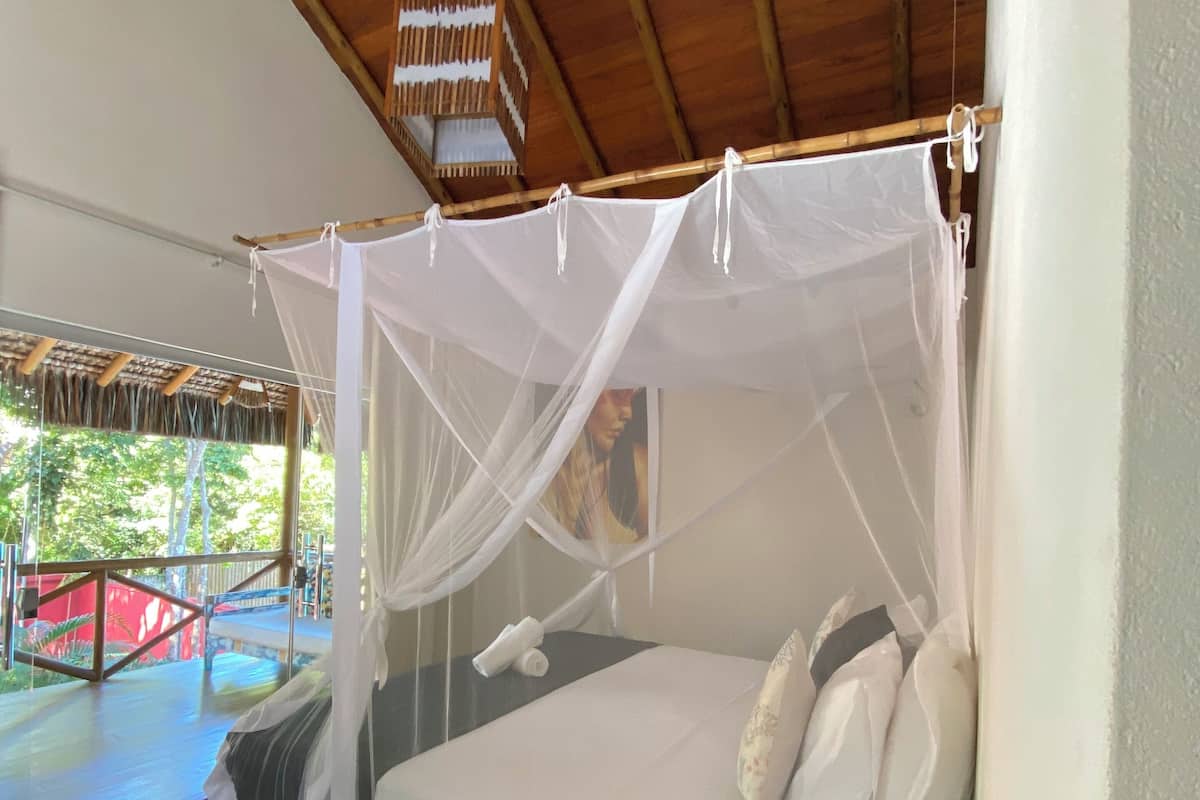 Una habitación con una cama bajo una mosquitera, un cabecero de madera, una mesita de noche con un control remoto y una ventana que da a una estructura roja.