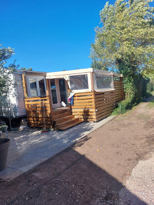 A small wooden cabin with a covered porch and a window. - Mobil Home 3 Ch, Grande Terrasse, un Camping Sécurisé, à 20mn à Pied de la Plage (Fréjus)