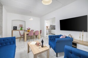 Un salon moderne avec un téléviseur à écran plat, des canapés bleus, une table à manger en bois et une cuisine en arrière-plan.