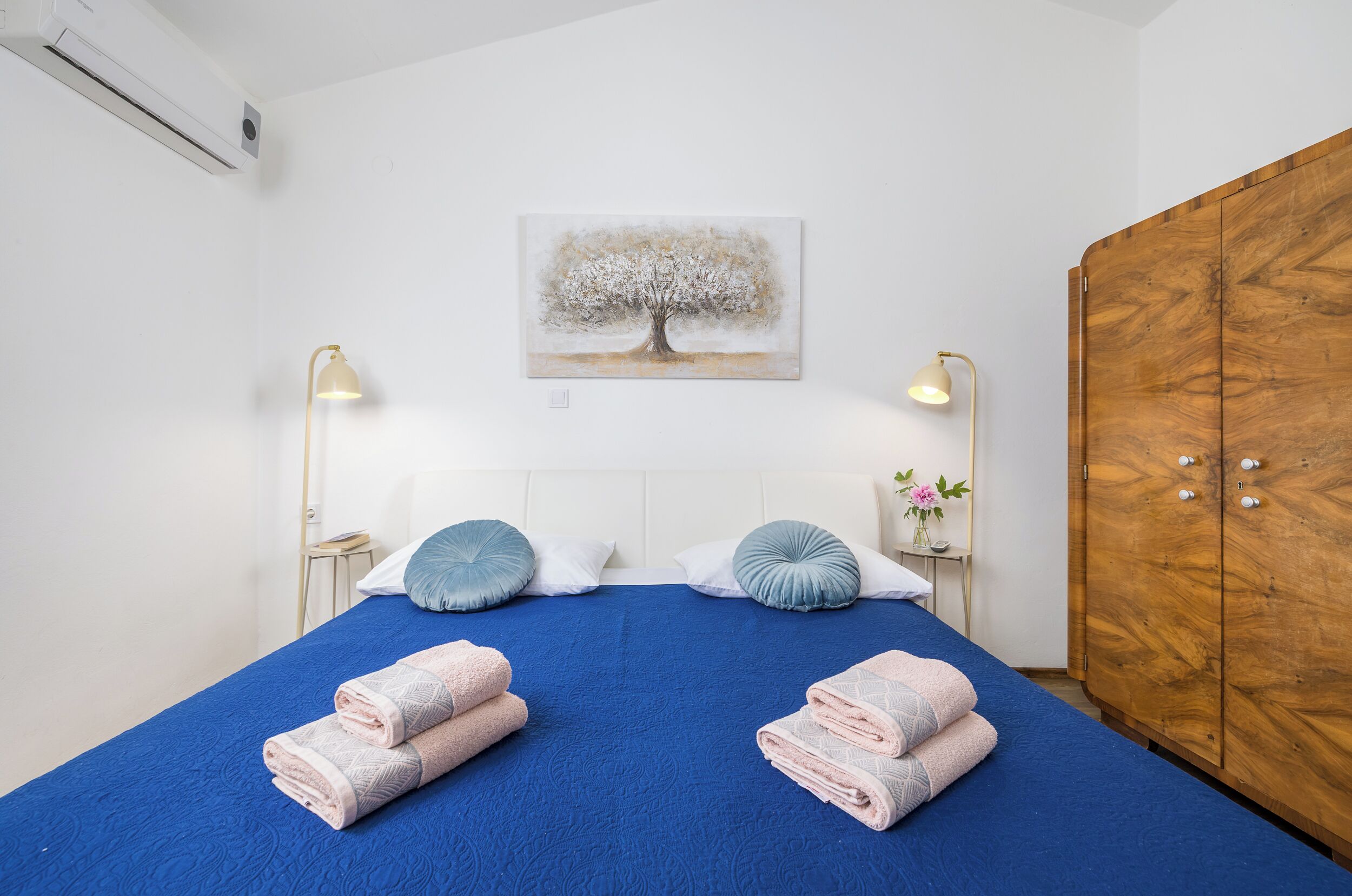 Ein ordentlich bezogenes Bett mit blauer Bettwäsche, zwei Nachttischlampen, ein Holzschrank und ein an der Wand hängendes Bild.