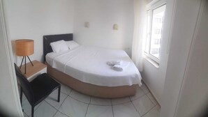 A small, neatly arranged bedroom with a bed, a bedside table, a lamp, and a chair. - Kibar Apart Pansiyon (Kusadasi)