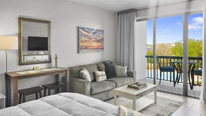 Une chambre d’hôtel moderne dotée d’un grand lit, d’un canapé, d’un bureau avec une télévision et d’un balcon offrant une vue sur le ciel et les arbres.