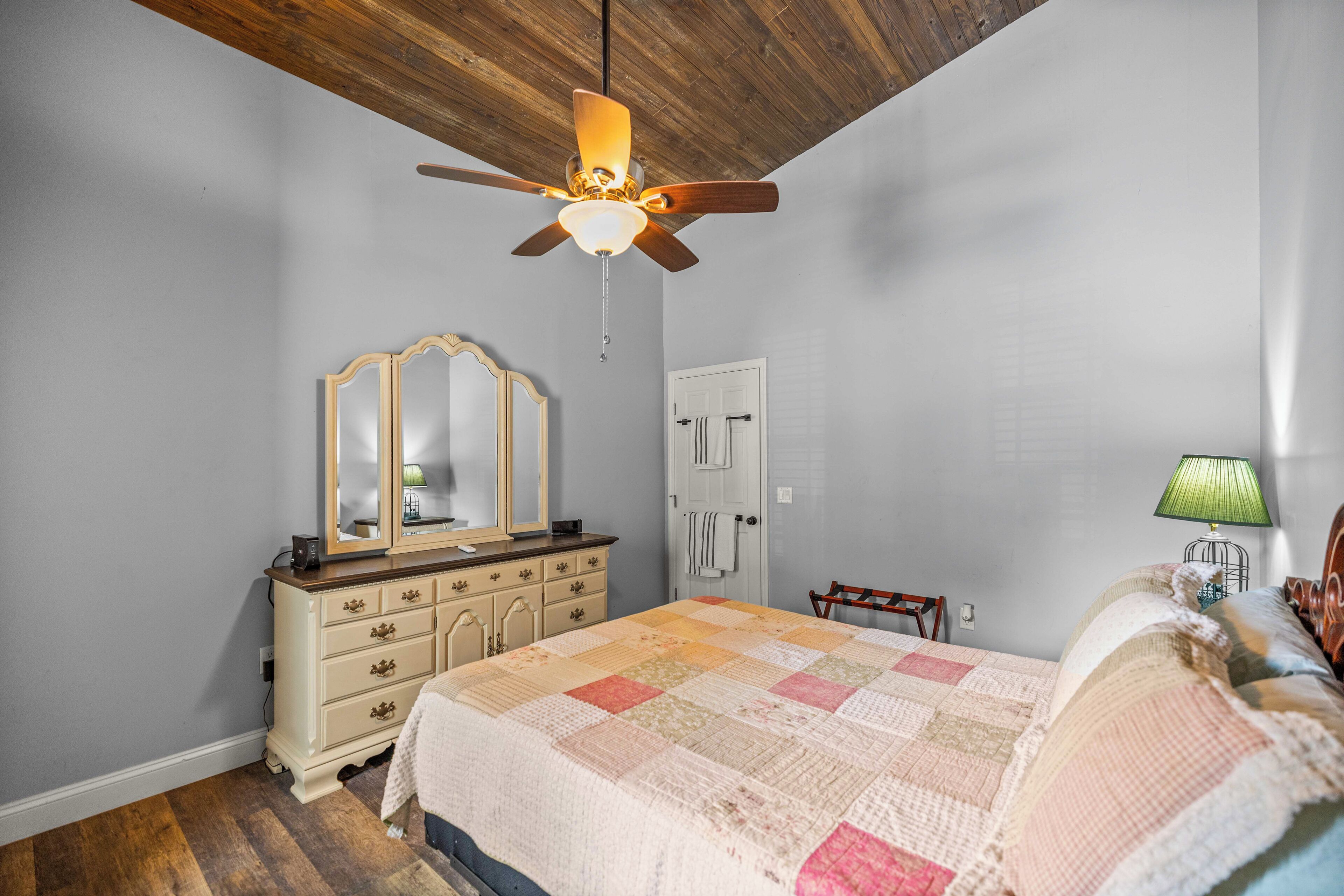 Un dormitorio con un ventilador de techo de madera, un aparador, una cama con colcha, un espejo y una puerta.