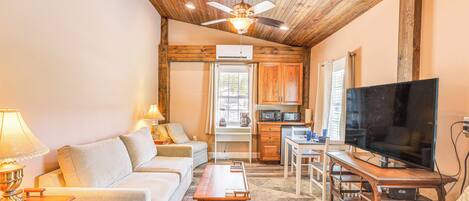 A cozy living room with a sofa, coffee table, and TV. There's a wooden ceiling fan, a kitchen area with cabinets, and a window with blinds.