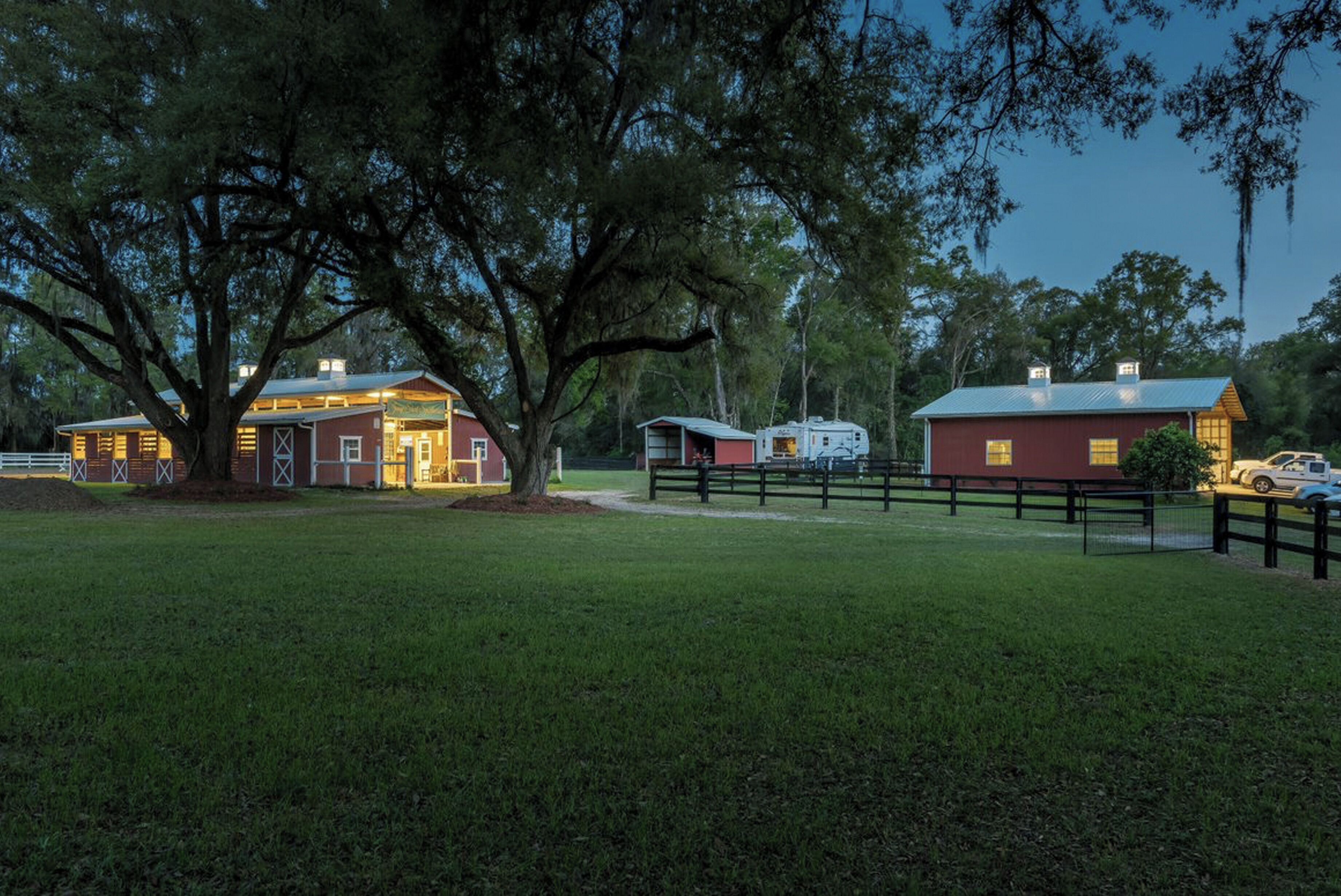 A large barn with a red exterior and a white roof, surrounded by a fenced area and a large tree.