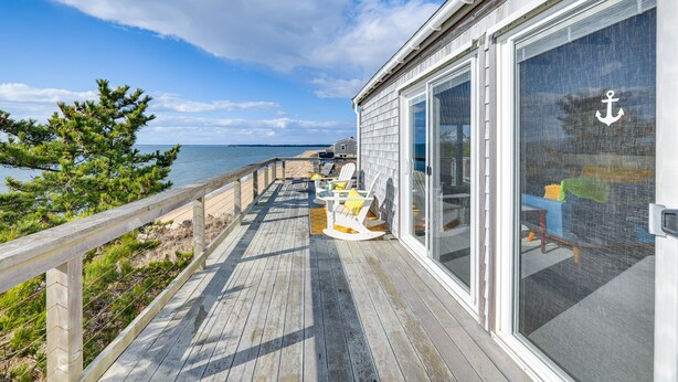 A wooden deck with outdoor seating, a view of the ocean, and a building with a large sliding glass door. - Eastham Beach House w/ Bay Views & Beach Access! (Eastham)