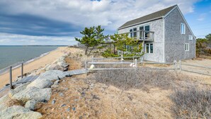 A coastal house with a balcony overlooking the beach, a wooden fence, and a rocky shoreline. - Eastham Beach House w/ Bay Views & Beach Access! (Eastham)
