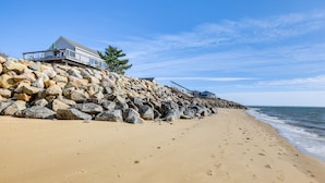 A beach with a rocky breakwater, a house with a balcony, and footprints in the sand. - Eastham Beach House w/ Bay Views & Beach Access! (Eastham)