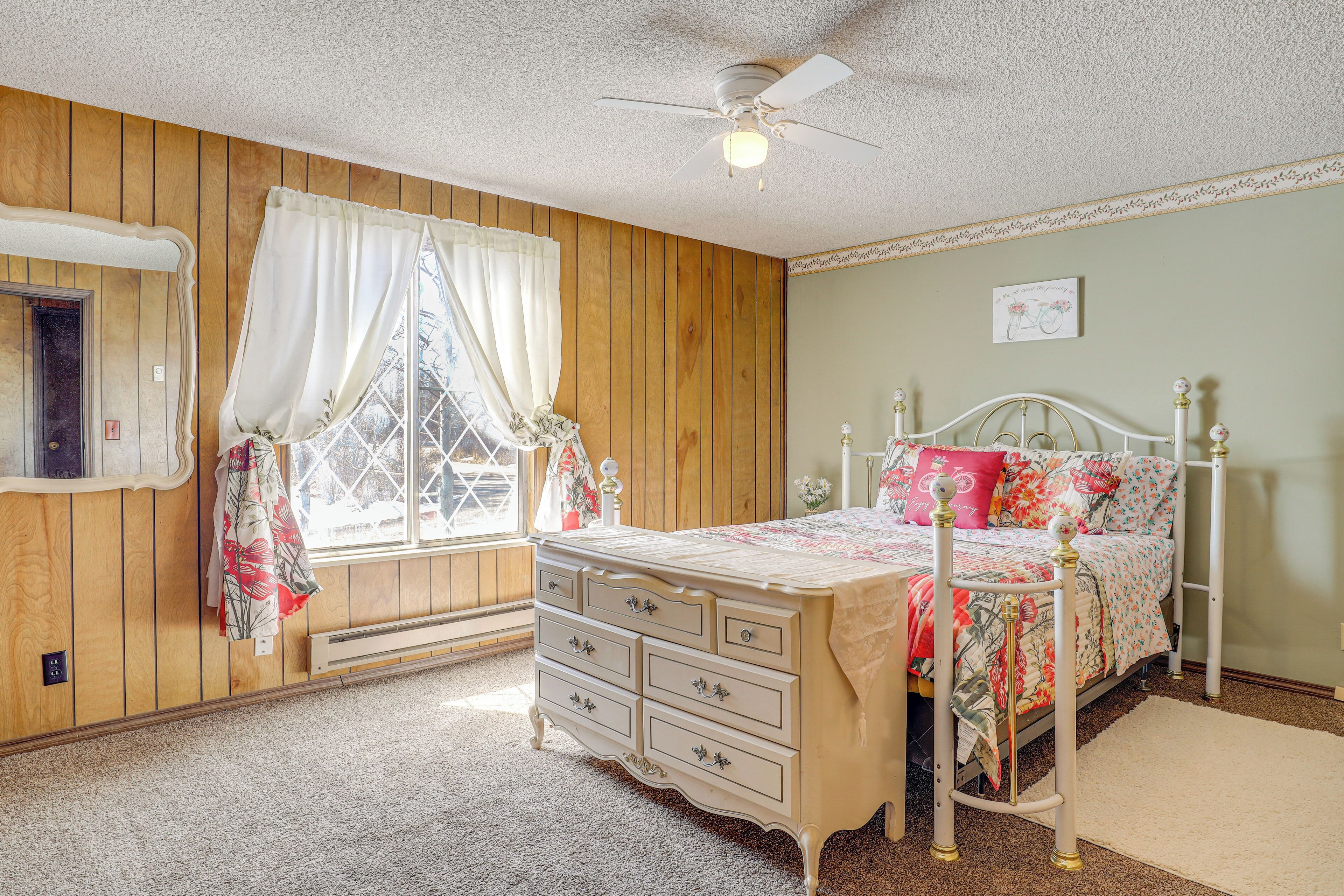 Een slaapkamer met een wand van houten panelen, een bed met een bloemenprint, een dressoir met laden, een plafondventilator en een raam met gordijnen.