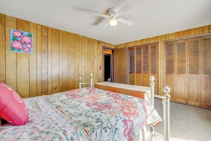 Une chambre avec des lambris en bois, un lit avec une literie à motifs floraux, un ventilateur de plafond et une armoire en bois.