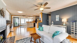 A spacious living room with a white sofa, a wooden coffee table, and a ceiling fan. A kitchen is visible in the background with a dining area. - Riverfront Colonial Beach Home w/ Private Beach! (Colonial Beach)