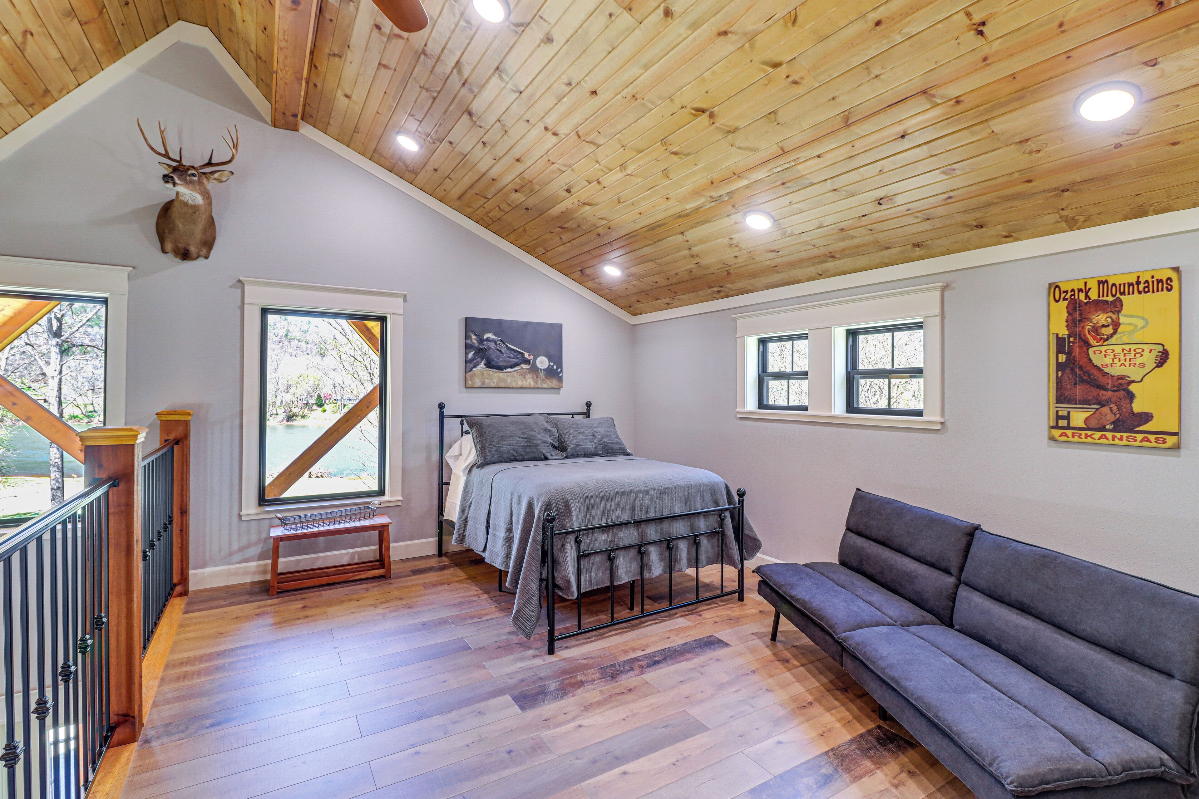 A bedroom with a bed, a gray sofa, a wooden ceiling, and a deer head mounted on the wall.