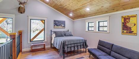 A bedroom with a bed, a gray sofa, a wooden ceiling, and a deer head mounted on the wall.