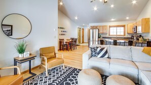 A spacious living area with a sofa, armchairs, a coffee table, and a dining table. There is a kitchen in the background with wooden cabinets and a refrigerator.