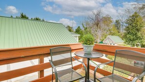 A balcony with two metal chairs and a small round table, a potted plant, and a view of a green-roofed building and trees.