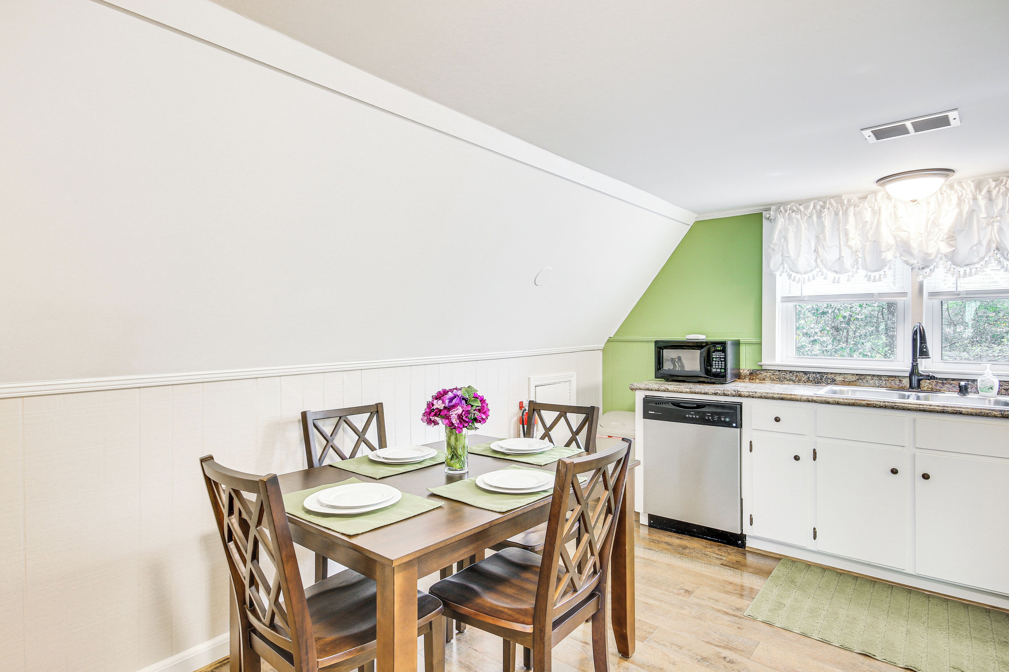 A dining area with a table set for four, a vase with flowers, and a kitchen with white cabinets and a green accent wall.