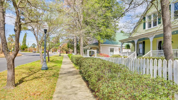 A residential street with a sidewalk, a white picket fence, and houses with green shutters.