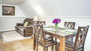 A dining area with a wooden table set for four, surrounded by wooden chairs. A brown leather sofa is positioned against the wall, and a vase with purple flowers is on the table.