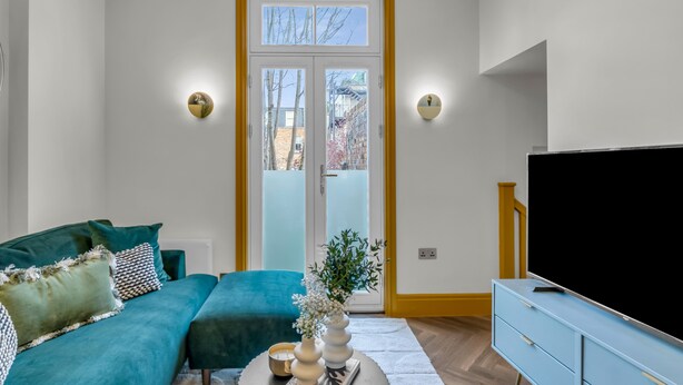 A modern living room with a teal sofa, a flat-screen TV, and a wooden cabinet. - Hammersmith Residences by MySquare (London)