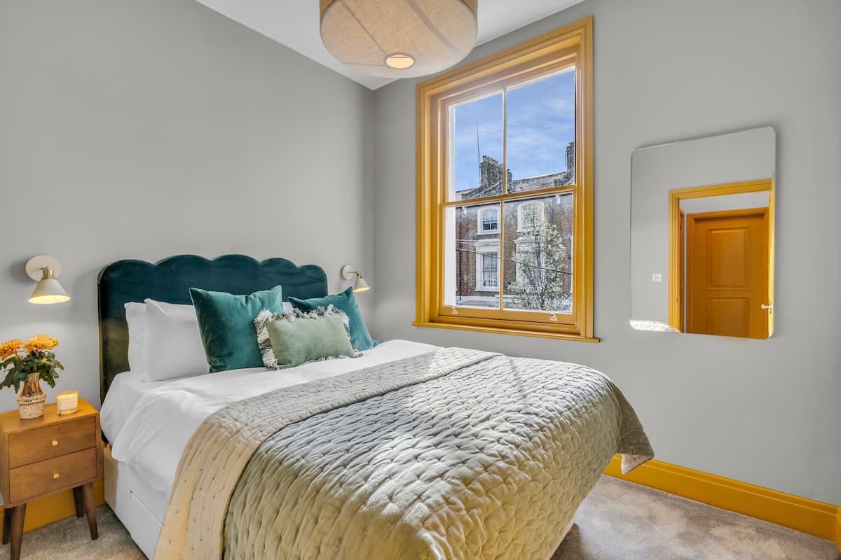 A bedroom with a large bed, a window with a view of buildings, a bedside table with a lamp, and a mirror on the wall.