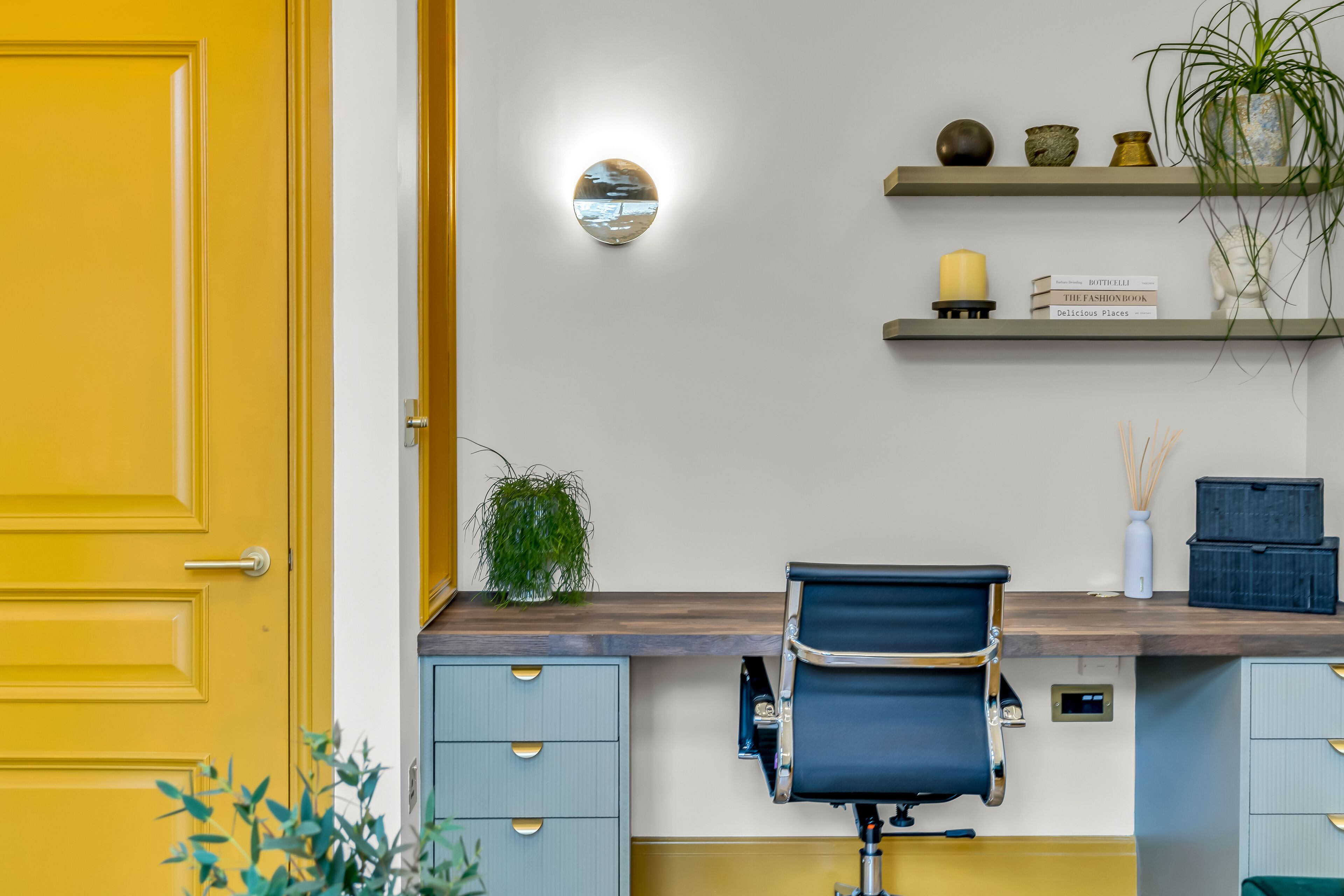 A modern office space with a yellow door, a desk with a chair, a round mirror, shelves with decorative items, and a potted plant.