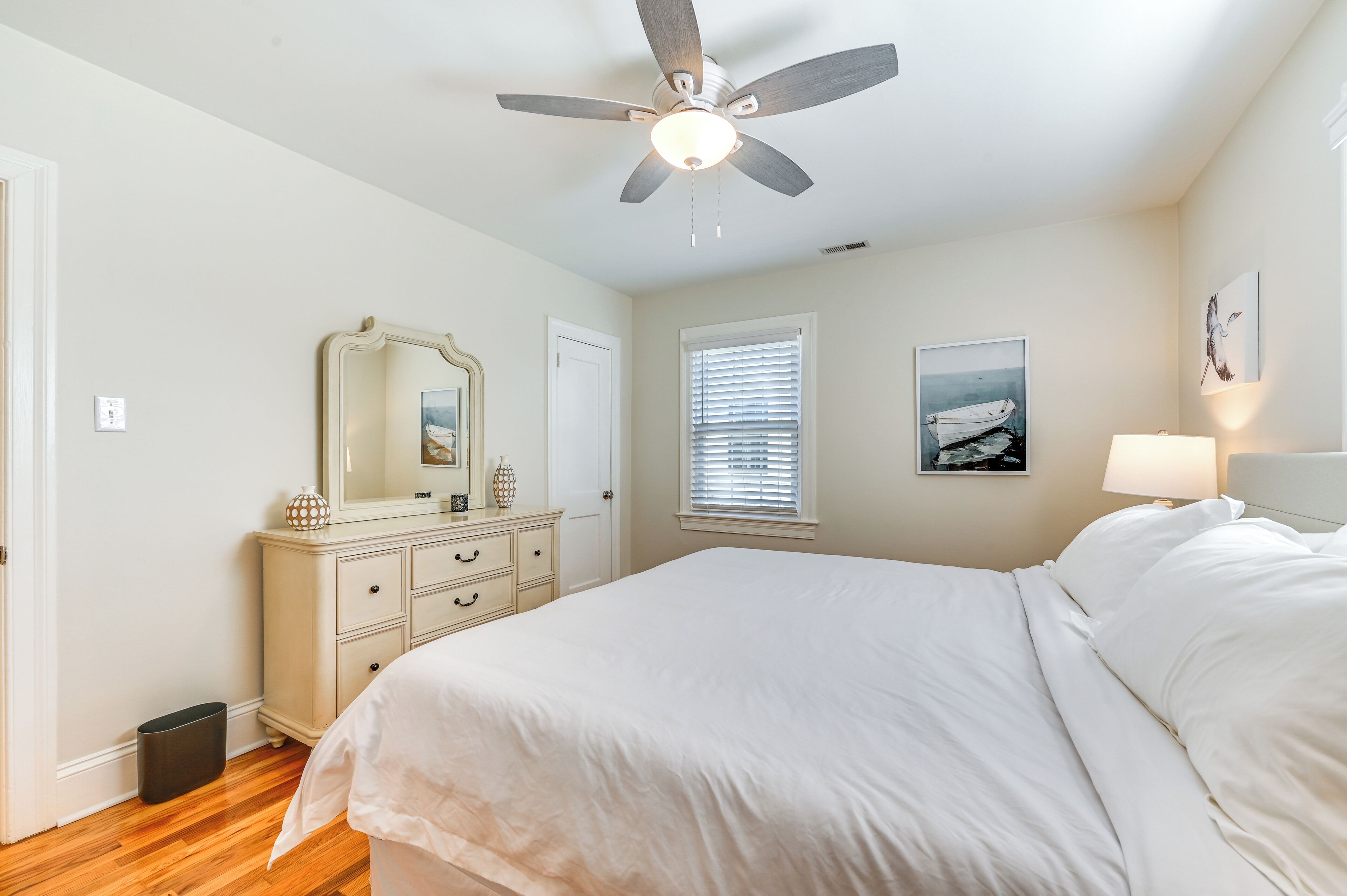 A bedroom with a bed, a dresser, a ceiling fan, a window with blinds, and a wall-mounted lamp.