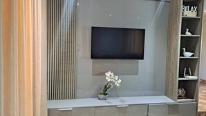 Ein modernes Wohnzimmer mit einem an der Wand befestigten Flachbildfernseher, einer kleinen Topf-Orchidee auf einem Konsolentisch und einer Holzregaleinheit mit Dekorationselementen.