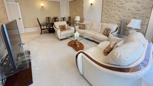 A living room with a sofa, armchairs, a glass coffee table, a TV, and a dining area with chairs and a table. - Tussauds Appartment (London)