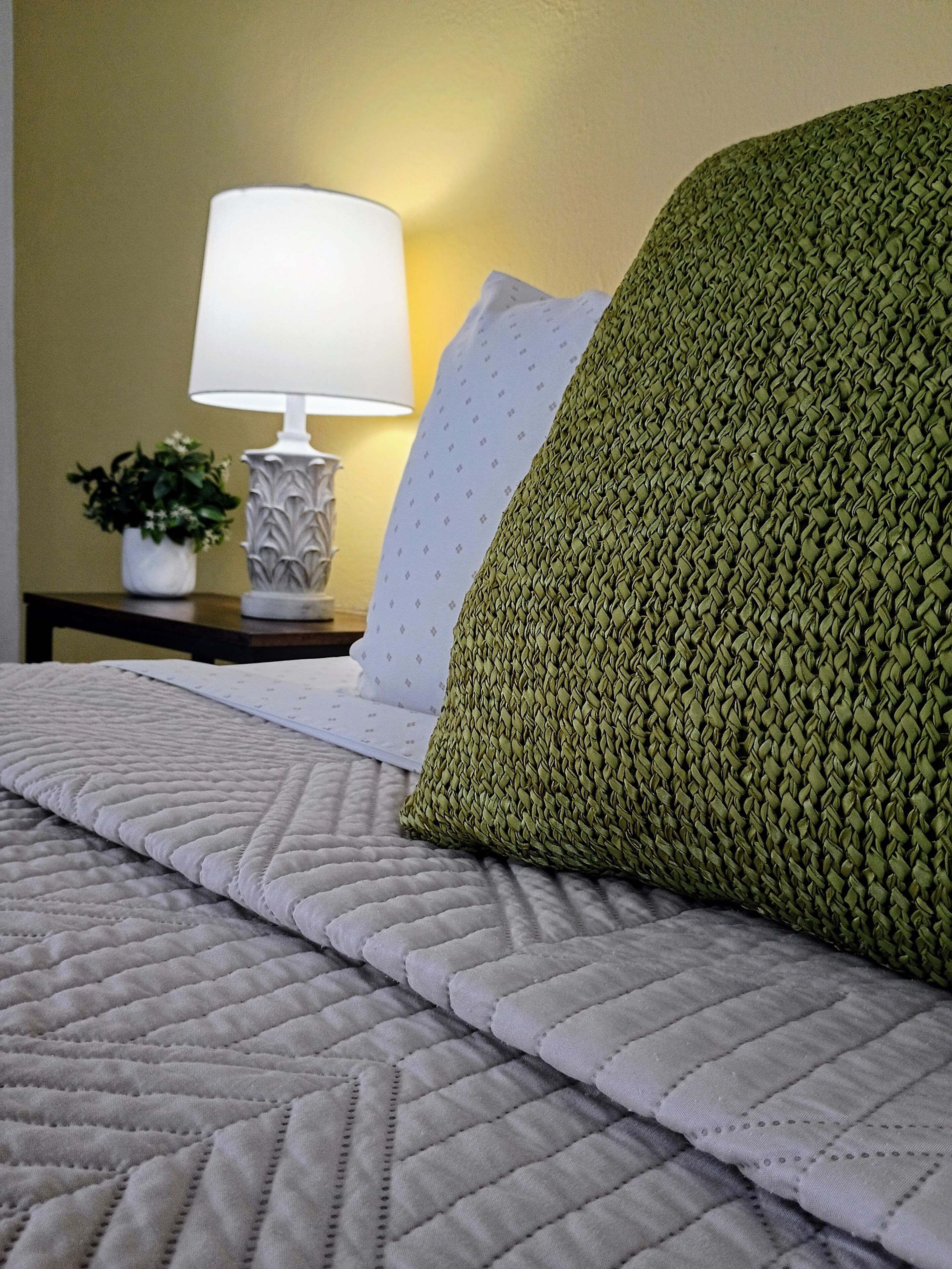 A bed with a quilted cover, a green knitted pillow, a white pillow, a bedside lamp, and a small potted plant on a nightstand.
