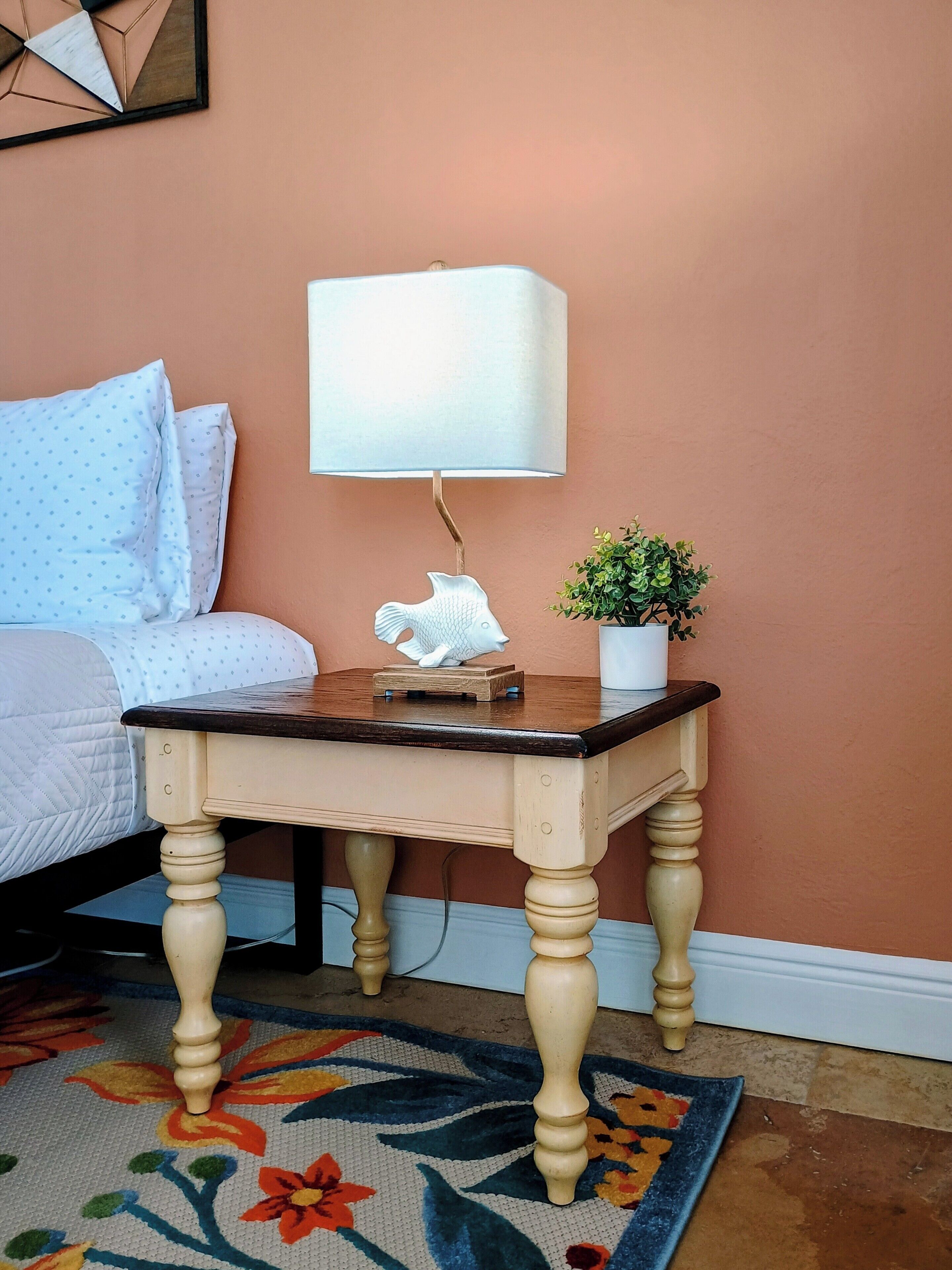 A bedroom with a bed, a nightstand with a lamp, a small plant, and a colorful rug.