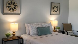 A neatly arranged bedroom with a bed, two bedside tables, two wall-mounted lamps, a chair, and two framed wall art pieces. - Bonsai - A Modern Motor Court (Rockledge)